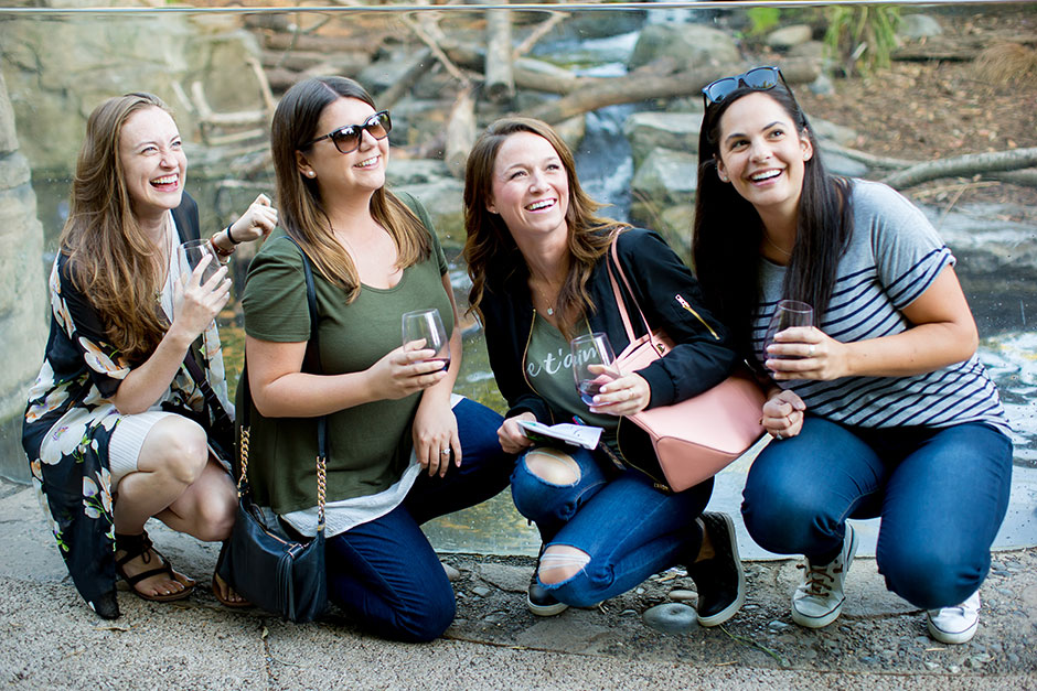 Four women crouch and smile while holding wine glasses outdoors. They are casually dressed and appear to be enjoying themselves, with trees and rocks visible in the background.