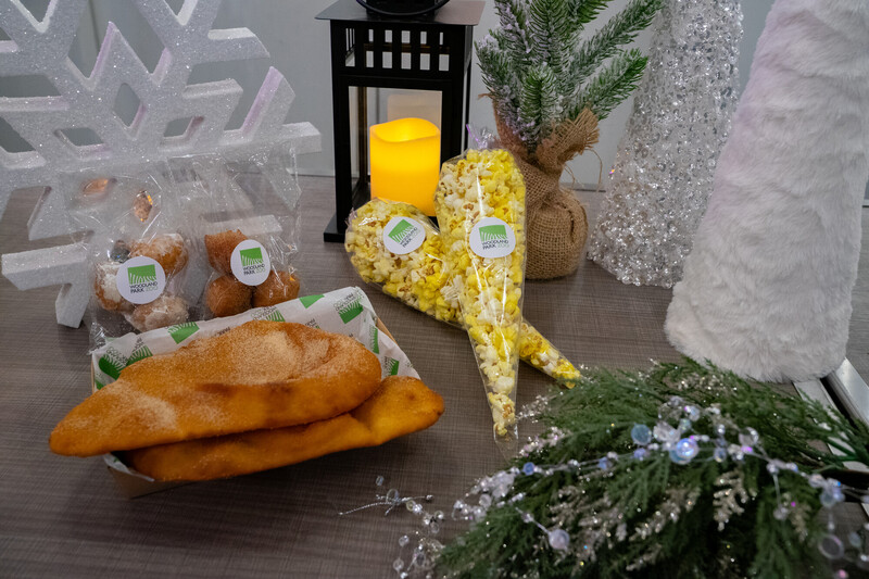 A festive table display with churros, powdered sugar doughnut holes, and a heart-shaped bag of popcorn, surrounded by holiday decorations like faux greenery, a lit candle, snowflake, and a sparkling Christmas tree.