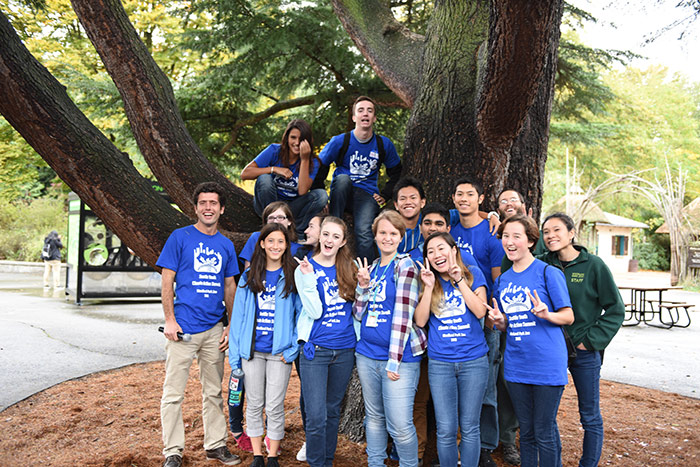 A group of smiling young people, most wearing matching blue t-shirts, pose together in front of a large tree outdoors. Some stand, while two sit on the tree branches. The setting appears to be a park or campus.