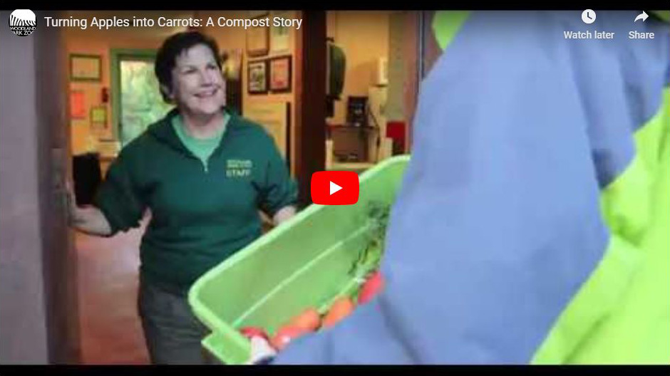 A woman smiling at a person holding a green bin filled with compostable food scraps, including carrots, at the entrance of a building. A YouTube video titled Turning Apples into Carrots: A Compost Story is shown.