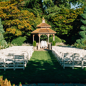 Một khung cảnh đám cưới ngoài trời với những hàng ghế trắng hướng về phía vọng lâu bằng gỗ, bao quanh là những hàng cây xanh tươi tốt dưới ánh nắng rực rỡ.