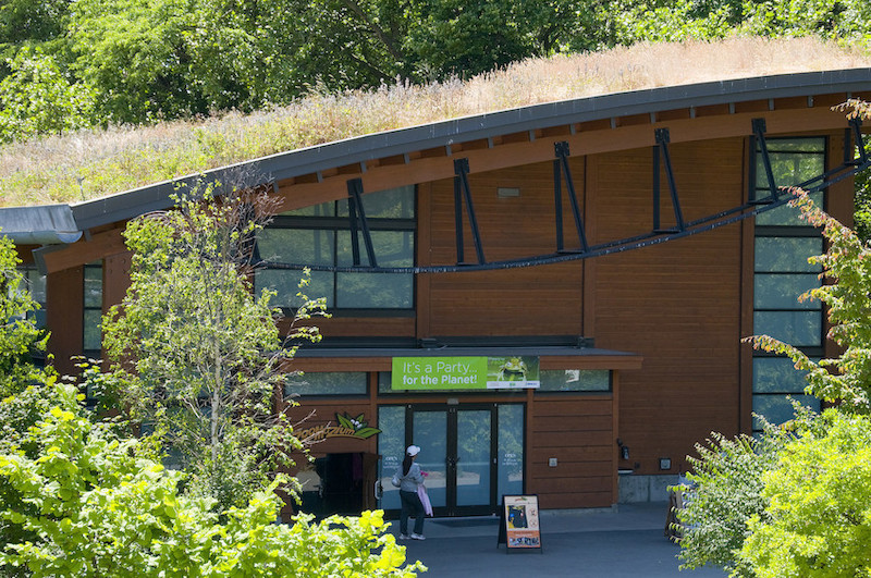 A modern building with a green, grass-covered roof is surrounded by lush trees. A person stands near the entrance, and a sign above the door reads, Its a Party for the Planet!.