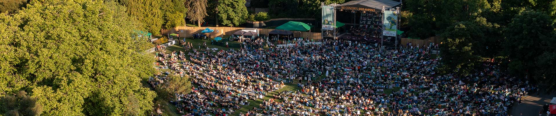 A large crowd gathers on a grassy lawn for an outdoor concert, sitting on blankets and chairs in front of a stage surrounded by trees and greenery on a sunny day.