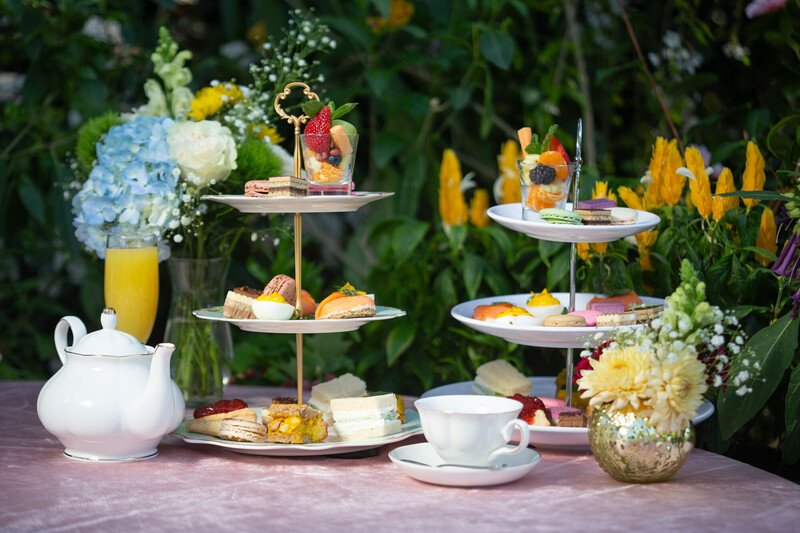 A table set outdoors with a white teapot, teacup, two tiers of assorted pastries and sweets, a glass of orange juice, and floral arrangements, all on a pink tablecloth with greenery in the background.