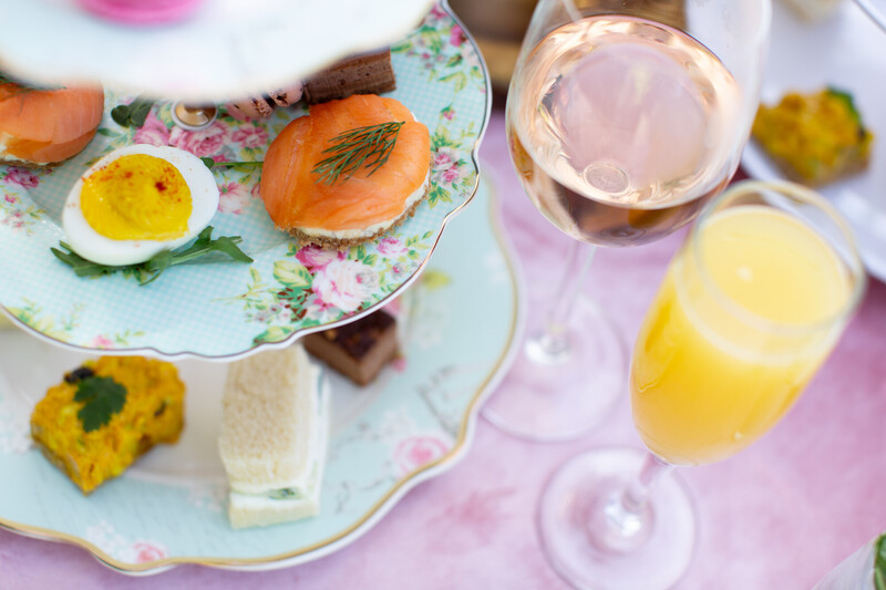 A close-up of an elegant afternoon tea setup with floral plates holding smoked salmon, a boiled egg, finger sandwiches, and pastries, alongside glasses of rosé wine and orange juice on a pink tablecloth.