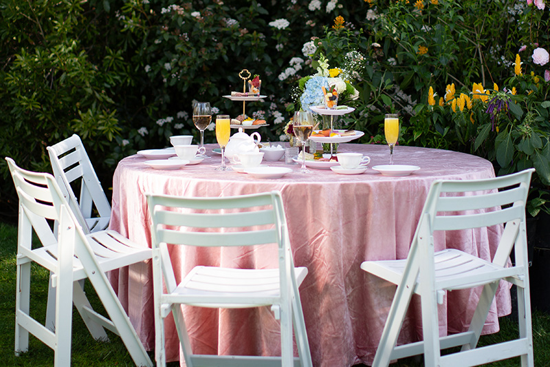 A round table covered with a pink tablecloth is set for tea in a garden. White chairs surround the table, which holds tiered trays of treats, teacups, wine glasses, and floral centerpieces. Lush greenery and flowers are in the background.