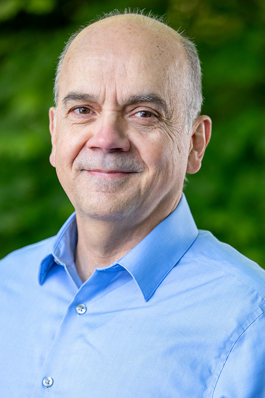 A middle-aged man with a receding hairline and light skin smiles softly. He is wearing a light blue collared shirt and is standing outdoors in front of blurred green foliage.