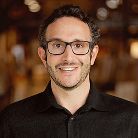 A man with short curly dark hair, glasses, and a trimmed beard smiles at the camera. He is wearing a black collared shirt and is standing in front of a warmly lit, blurred background.