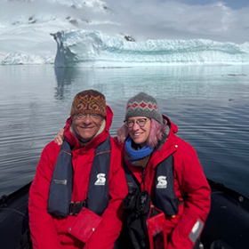Two people in red jackets and winter hats sit together on a boat in icy waters, smiling at the camera with a large glacier and snow-covered mountains in the background.