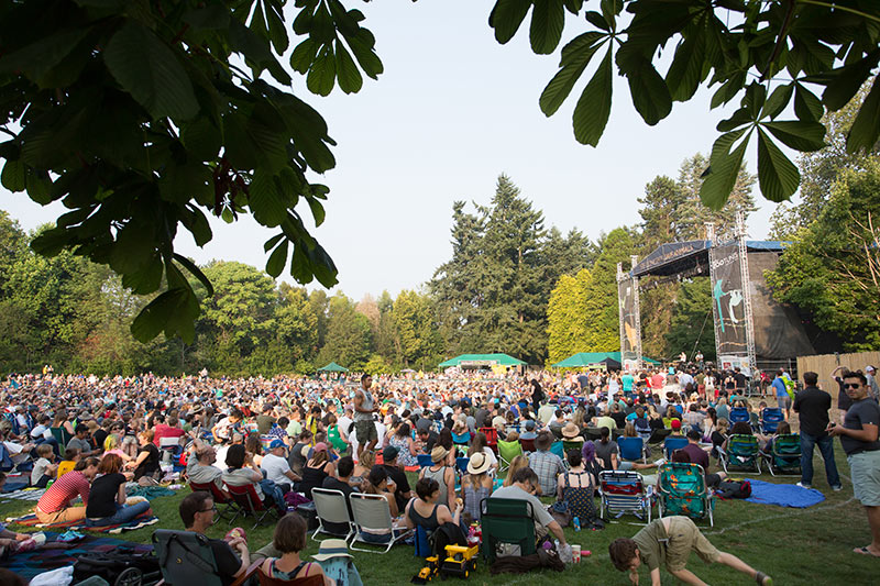 A large crowd gathered outdoors at a park music festival, many sitting on blankets and lawn chairs, facing a stage surrounded by trees under a clear sky.