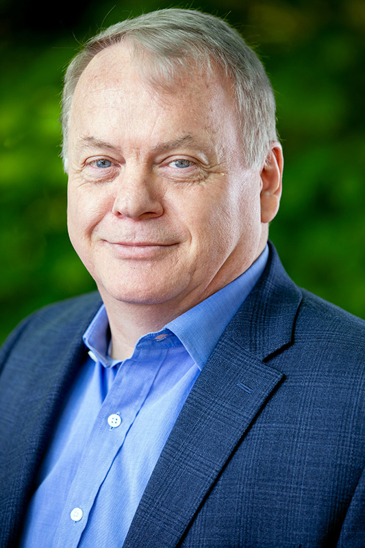 A middle-aged man with short gray hair, wearing a blue dress shirt and dark blazer, smiles softly at the camera. The background is blurred with green foliage.