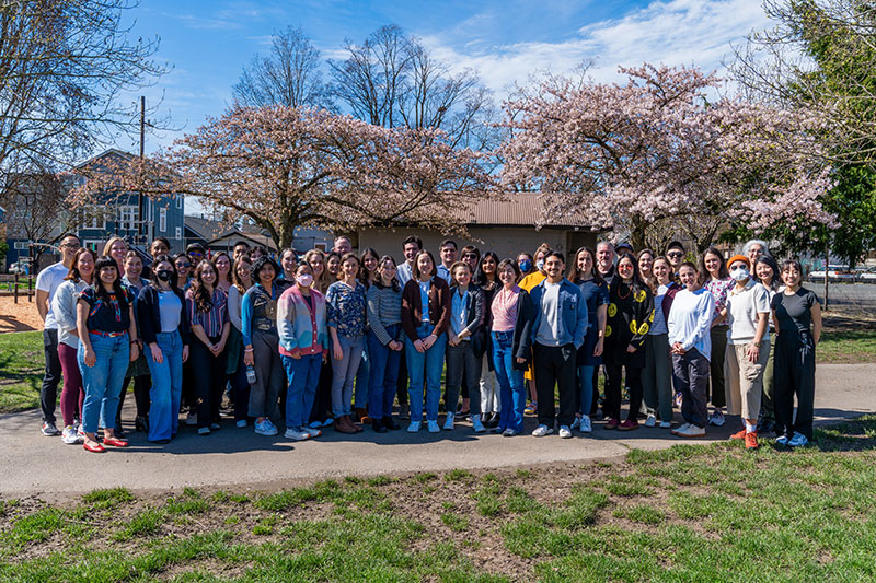 A large group of people stands together outdoors in a park, posing for a photo in front of blossoming trees and a small building on a sunny day.