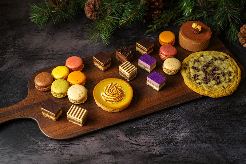 A wooden board topped with assorted desserts, including colorful macarons, layered pastries, a lemon tart, a chocolate cookie with chips, a chocolate cake, and festive greenery in the background.