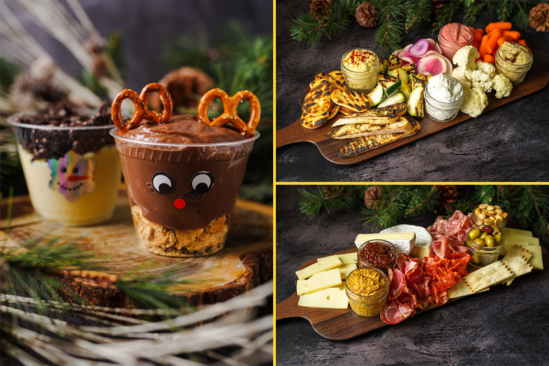 A festive dessert cup decorated as a reindeer, next to a snowman cup, sits on a wooden slab; beside them are two boards: one with assorted hummus and veggies, the other with cured meats, cheese, and nuts. Pine branches decorate the scene.