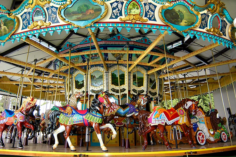 一座色彩斑斓的复古旋转木马，顶棚上有精美的彩绘马匹和装饰艺术品，矗立在金属屋顶下，等待游乐园的乘客。