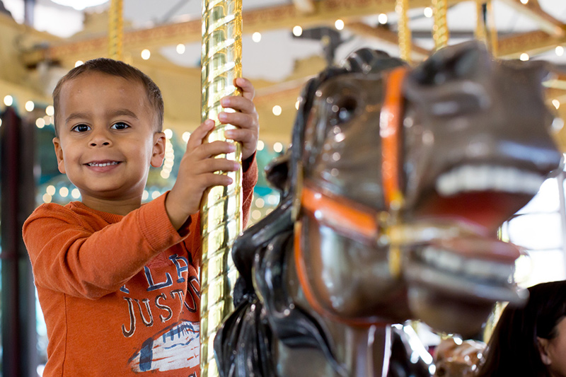 Un niño con camisa naranja sonríe mientras monta un caballo de carrusel. El fondo está decorado con guirnaldas de luces, creando un ambiente festivo. 