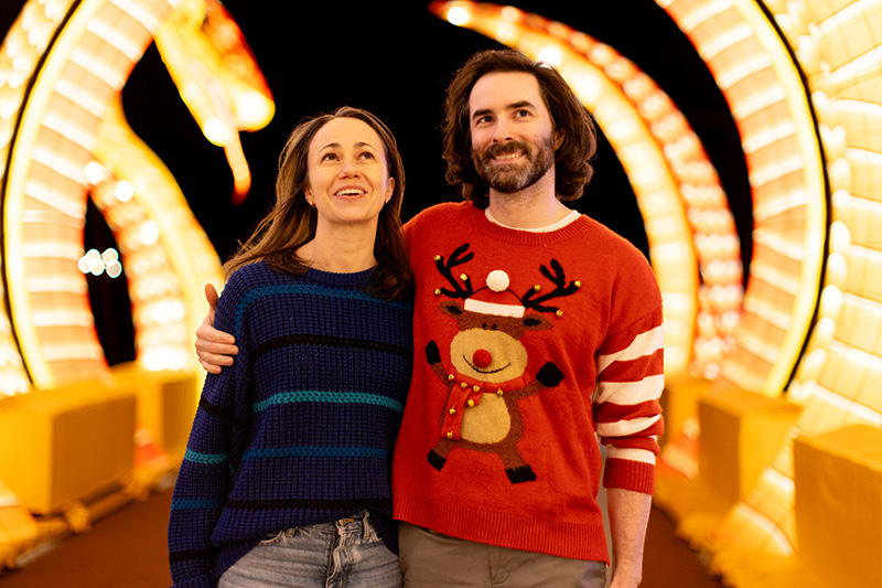 A smiling couple stands arm-in-arm at night in front of a brightly lit dragon display. The woman wears a blue striped sweater, and the man wears a red sweater with a reindeer design. Both look happy and festive.