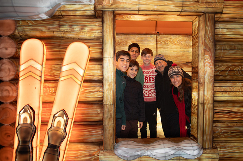 Six people, including four boys and two adults, smile while standing inside a wooden cabin. Decorative skis and boots are mounted outside the window where they are gathered. Everyone wears winter clothing.