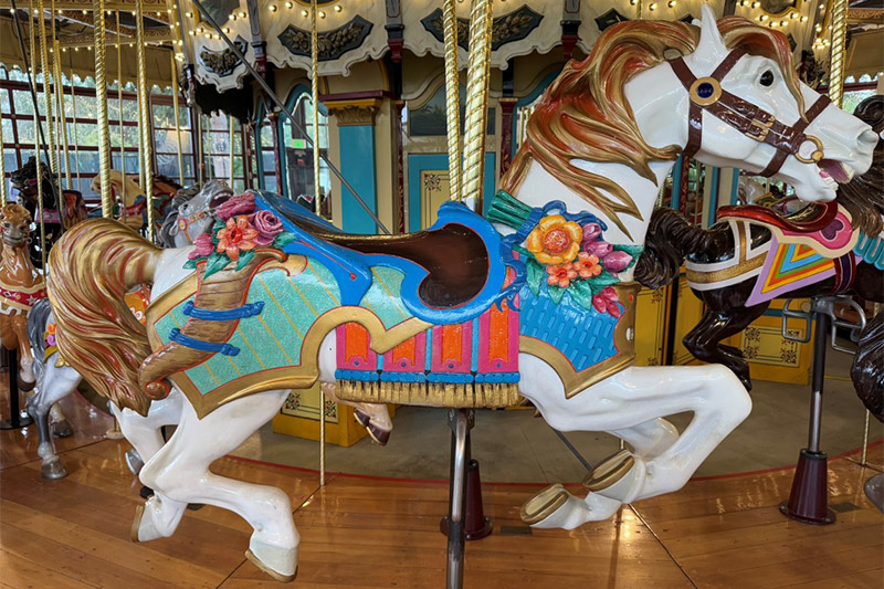 A close-up of a colorful carousel horse, decorated with flowers and a bright blue, red, and gold saddle. The horse is white with a golden mane and tail, set on a shiny wooden platform.