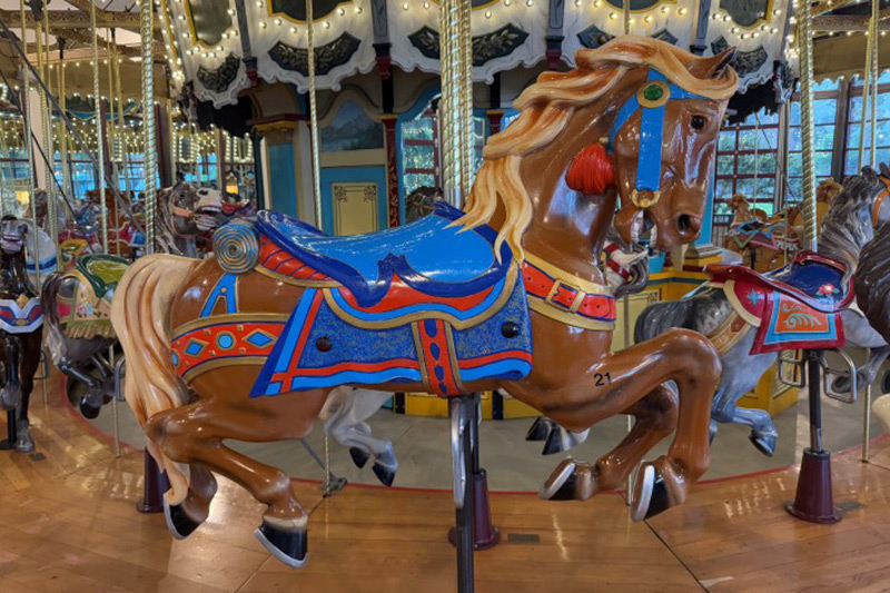 A brightly painted carousel horse with a blue, red, and orange saddle and bridle, mounted on a classic indoor carousel surrounded by other colorful horses and lights.