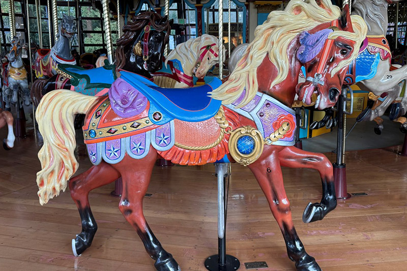 A brightly painted carousel horse with a blue saddle, decorative ornaments, and flowing blonde mane stands on a wooden floor among other carousel horses.