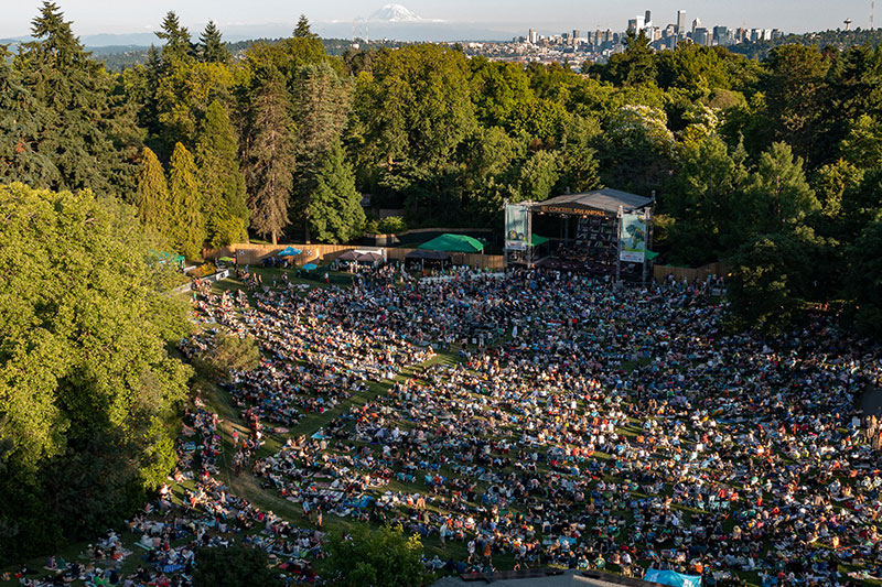 A large crowd gathers on a grassy hillside for an outdoor concert, surrounded by tall trees, with a city skyline and a snow-capped mountain visible in the distance under a clear sky.