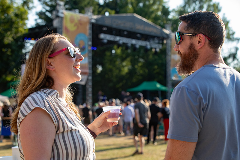 A woman and a man in sunglasses smile and talk at an outdoor music festival, with a cup in the womans hand and a crowd gathered near a stage in the background.