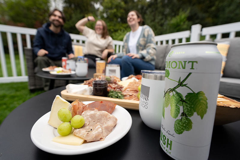 Three people sit and smile on patio furniture outdoors, with a table in the foreground holding a can of beer, a plate of deli meats, cheese, grapes, and other snacks. A white fence and trees are in the background.