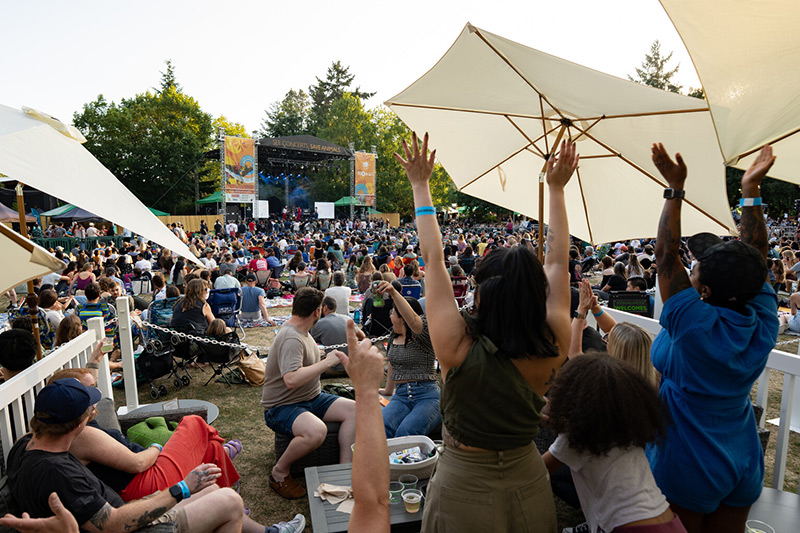 A large crowd enjoys an outdoor concert, with people sitting on the grass and standing, some with arms raised. Trees and large umbrellas provide shade, and a band performs on a stage in the background.