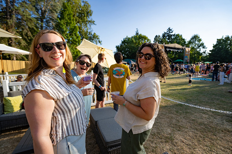 Three women wearing sunglasses smile at the camera while holding drinks at an outdoor event on a sunny day. People, trees, and a stage are visible in the background.