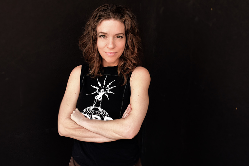 A woman with wavy brown hair stands against a dark background, arms crossed, wearing a sleeveless black shirt featuring a white graphic design of a person inside a starburst.