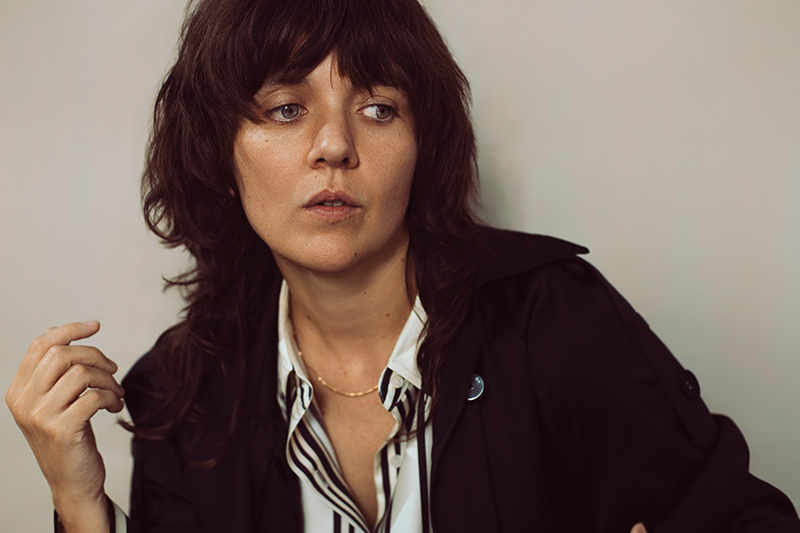 A person with shaggy dark brown hair and bangs wears a black coat over a striped shirt, looking slightly to the side with a thoughtful expression against a plain background.