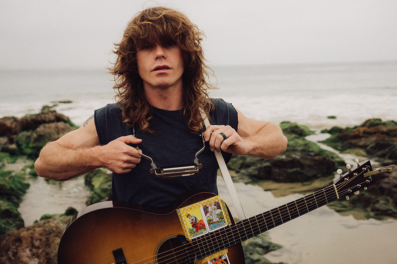 A person with long hair sits on the grass in a sunlit forest, holding an acoustic guitar. They wear a white t-shirt and jeans, gazing thoughtfully at the camera.