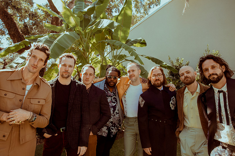 Eight men stand closely together outdoors in front of lush green plants and a white wall, dressed in stylish, semi-formal clothing. The group smiles and poses in the sunlight.