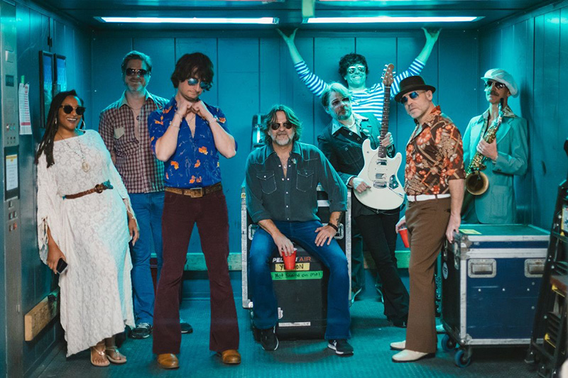 A group of nine adults in retro, colorful 1970s-style clothing pose confidently in an elevator. Some hold musical instruments, including a guitar and saxophone, suggesting they are a band. Everyone looks relaxed and playful.