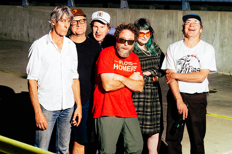 Six people stand together outdoors in bright sunlight, posing casually. One person is wearing a red shirt and sunglasses, another has green hair and orange glasses, and the group appears relaxed and smiling in front of a concrete wall.