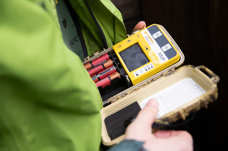 A person in a green jacket holds an open yellow detector device with visible batteries and a screen, next to a small plastic case containing a paper insert.
