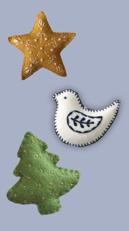 Three felt ornaments on a gray background: a brown star with white stitching, a white bird with blue embroidery, and a green Christmas tree with small gold decorations.