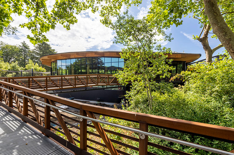 A modern, circular building with large glass windows is surrounded by lush green trees. A wooden bridge with metal railings leads to the building, blending into the natural landscape under a partly cloudy sky.