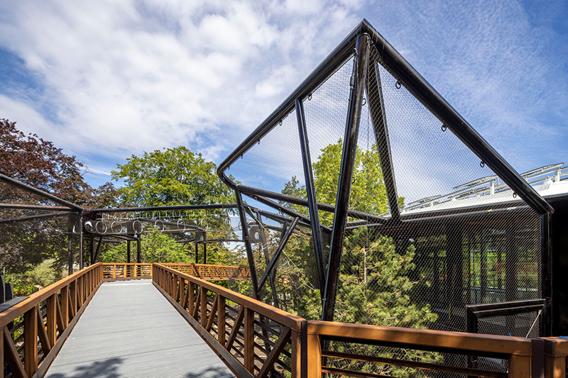 Modern pedestrian bridge with wooden railings and black metal framework, set among green trees under a partly cloudy sky. The bridge connects to a glass building on the right.