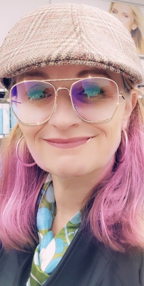 A person with pink hair, wearing a flat cap, large wire-frame glasses, and hoop earrings, smiles at the camera. They have a green and blue scarf, and there is a blurred background with a face visible on a poster.