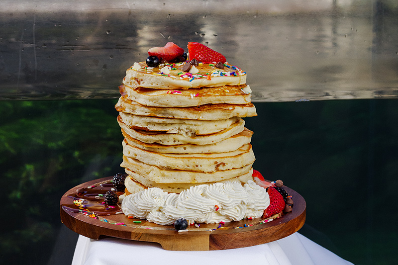 A tall stack of pancakes topped with berries and sprinkles, surrounded by whipped cream, berries, and chocolate on a wooden platter. The background is blurred.