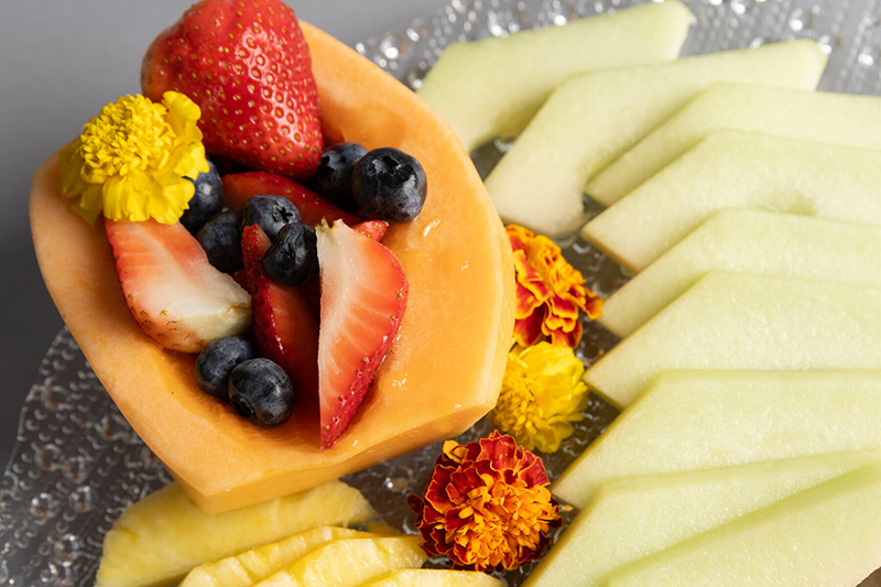 A clear plate with sliced honeydew melon, pineapple, and a cantaloupe bowl filled with strawberries and blueberries, garnished with yellow and orange edible flowers.