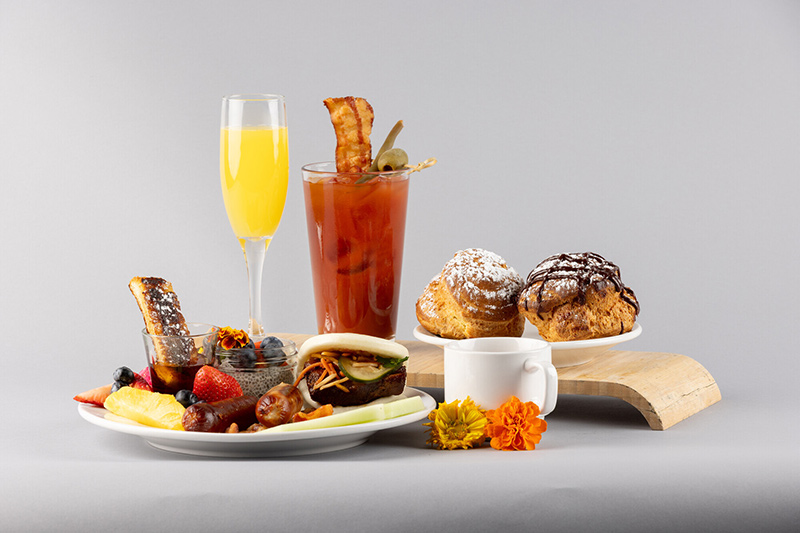 A brunch spread featuring a mimosa, a Bloody Mary with bacon and olives, a plate with fruit, toast, and a sandwich, two cream puffs dusted with powdered sugar, a white coffee cup, and orange flowers on a light background.