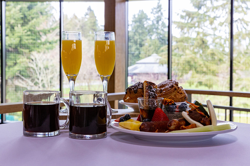 A breakfast table with two mugs of coffee, two glasses of orange juice, a plate of assorted pastries, and a plate of fruit, sausage, and vegetables, set by large windows overlooking trees.