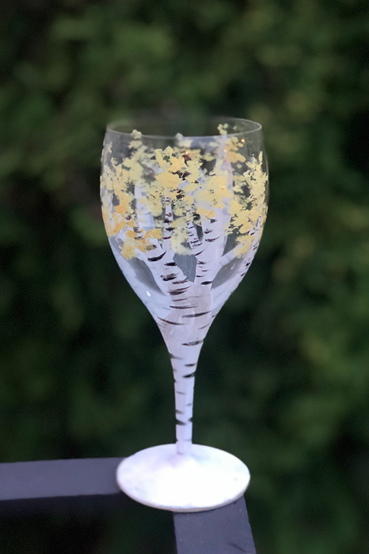A wine glass hand-painted with white birch trees and yellow autumn leaves, set against a blurred green background.