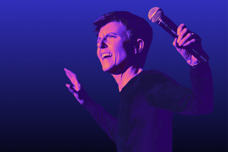 A person holding a microphone gestures expressively while performing on stage, lit with dramatic blue and purple lighting.