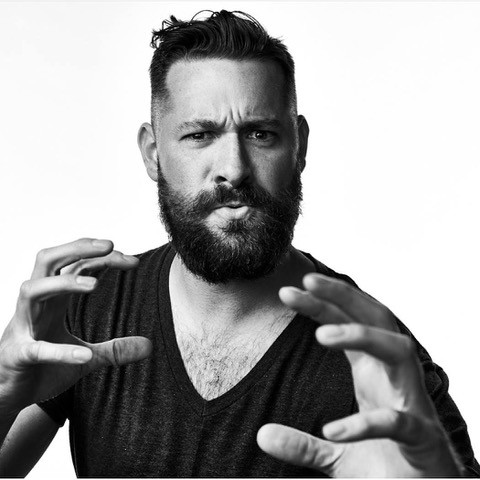 A bearded man with short, styled hair and a serious expression gestures with his hands as if emphasizing a point, posing against a plain white background.