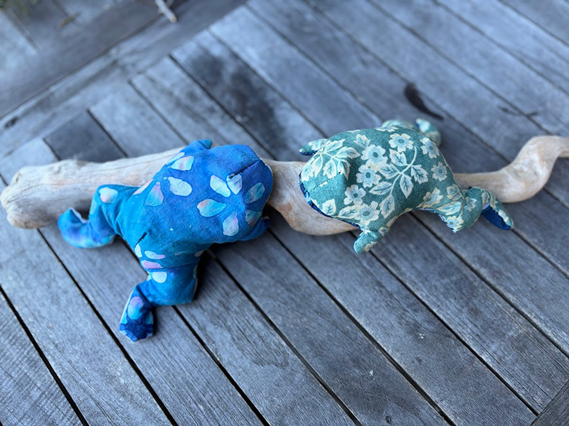 Two fabric frogs, one blue with light spots and the other green with a floral pattern, rest on a piece of driftwood on a weathered wooden surface.