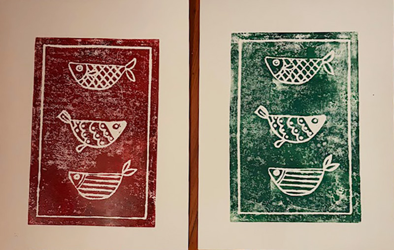 Two prints side by side show three stylized fish in a rectangular border. The left print is red and the right print is green, both with a similar simple, hand-carved design and some uneven ink distribution.